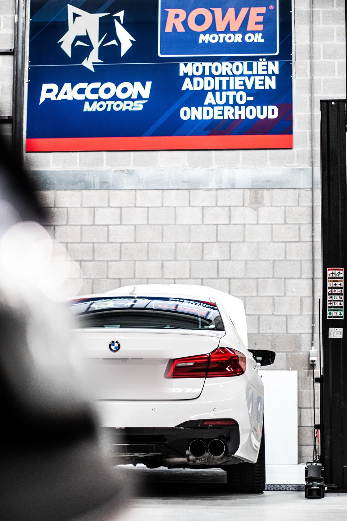 Een witte BMW M5 in de werkplaats, onder het bord van Raccoon Motors en ROWE Motor Oil