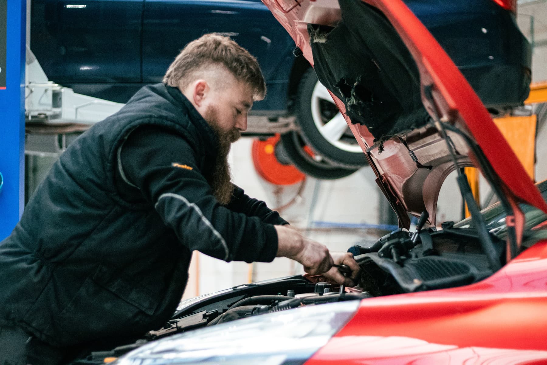 Technieker werkt in de motorruimte van een rode Renault Clio, met een wagen op de hefbrug erachter