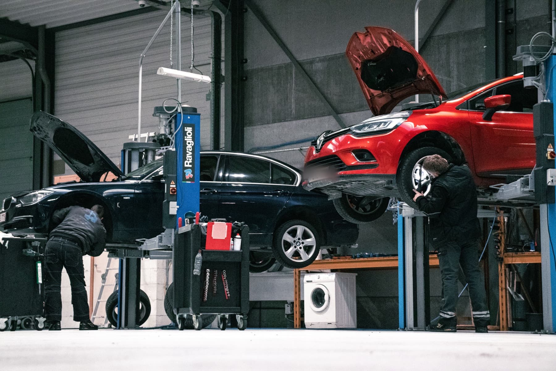 Twee techniekers in de werkplaats: een donkerblauwe BMW 3-serie met open motorkap en een rode Renault Clio op de hefbrug